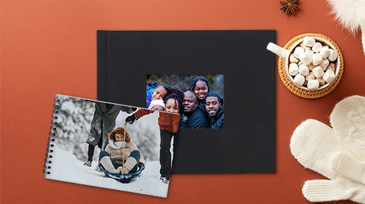 Same Day photo books: A black hardcover photo book with a family photo inset and an open spiral-bound photo book displaying a winter snow photo, placed on a red background with hot cocoa and white mittens.
