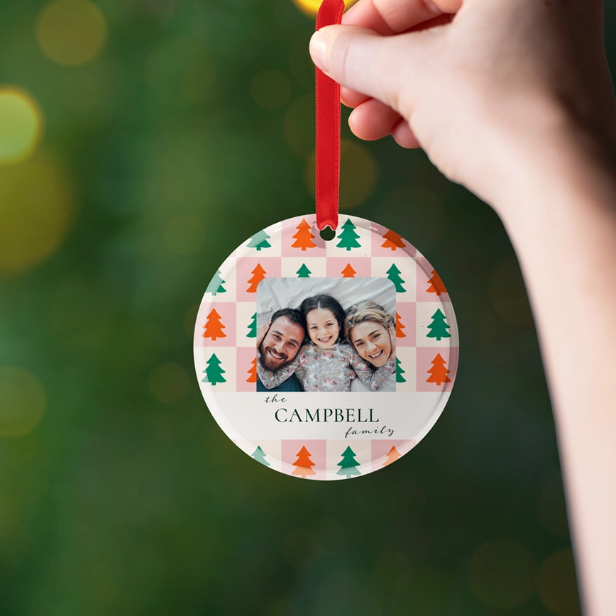Circle acrylic ornament: Round glossy acrylic ornament with a red ribbon, featuring a checkerboard pattern of pink and cream squares with orange and green Christmas trees, a photo of a woman kissing a child, and the text the Calderon family.