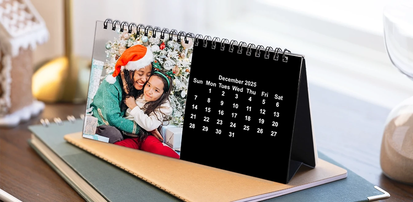 A personalized photo desk calendar shown on an office desk, featuring the month December 2025 and a photo of two smiling people, one wearing a Santa hat, embracing in front of a decorated Christmas tree.