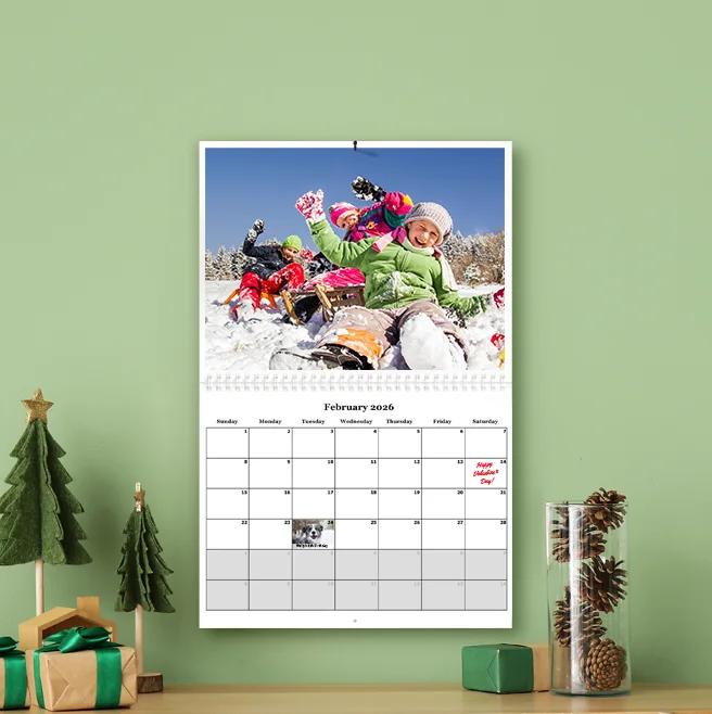 Photo calendar, A personalized wall calendar for February 2026 hanging on a green wall, featuring a large photo of children playing in the snow and a small pet photo inset on the date grid.