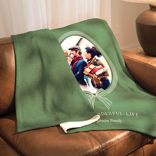 Shop blankets: A green plush fleece photo blanket folded over a brown leather couch, featuring a photo of a family holding presents within a white oval frame, and the text WONDERFUL LIFE Johnson Family.