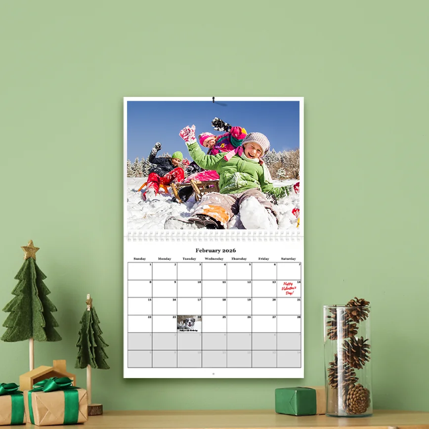 Photo calendar, A personalized wall calendar for February 2026 hanging on a green wall, featuring a large photo of children playing in the snow and a small pet photo inset on the date grid.