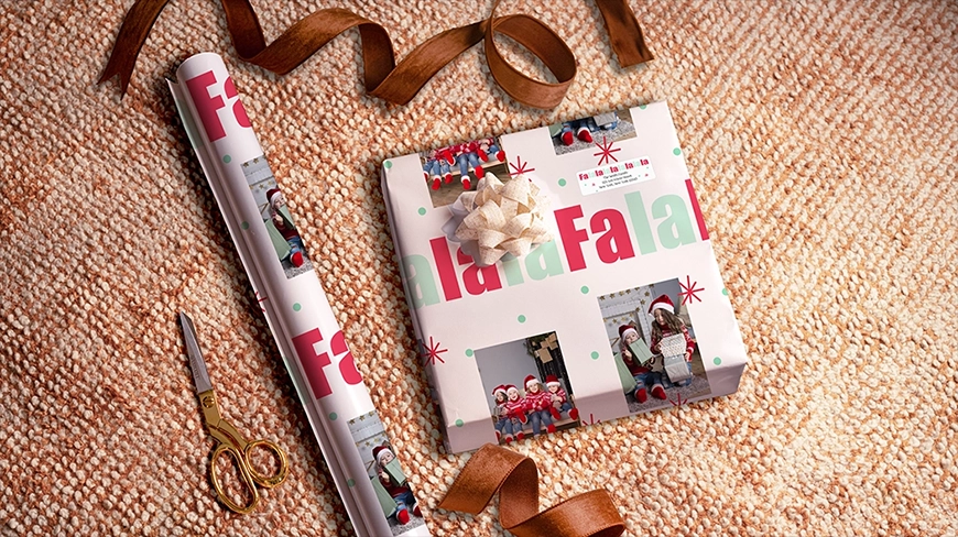 A festive holiday scene showing a roll of custom photo wrapping paper next to a wrapped gift, both featuring a Fa la la design with photos of children in elf costumes. The gift, wrapped with the custom paper and a beige bow, is on a textured brown surface with scissors and brown ribbon nearby.